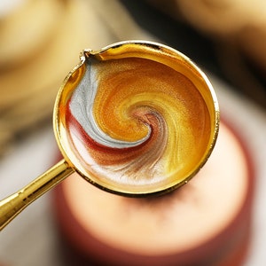 May include: A gold-toned metal spoon with a round, shallow bowl filled with a swirling mixture of gold, silver, and red wax.