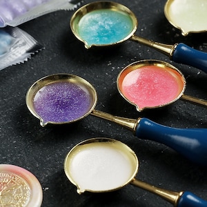 May include: Four gold-toned wax seal spoons with blue, purple, pink, and white wax. The spoons have dark blue handles.