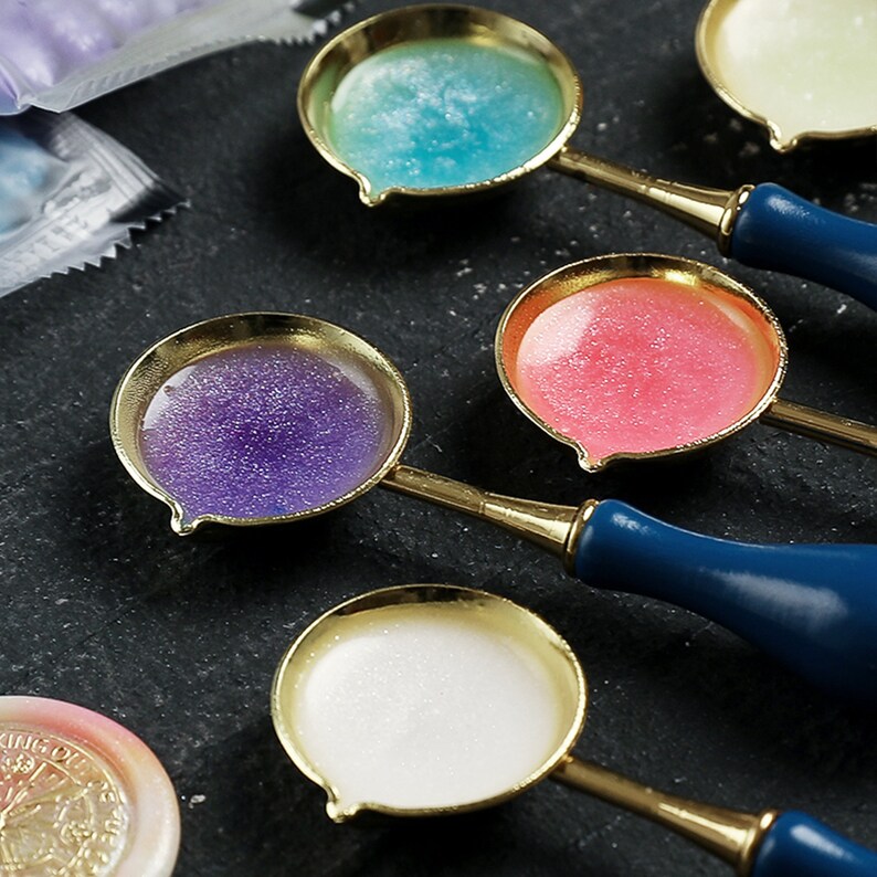 May include: Four gold-toned wax seal spoons with blue, purple, pink, and white wax. The spoons have dark blue handles.