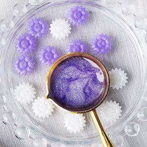 May include: A small glass dish with a gold rim contains a variety of purple and white flower-shaped resin embellishments. A small gold spoon with a purple glittery liquid sits in the center of the dish.