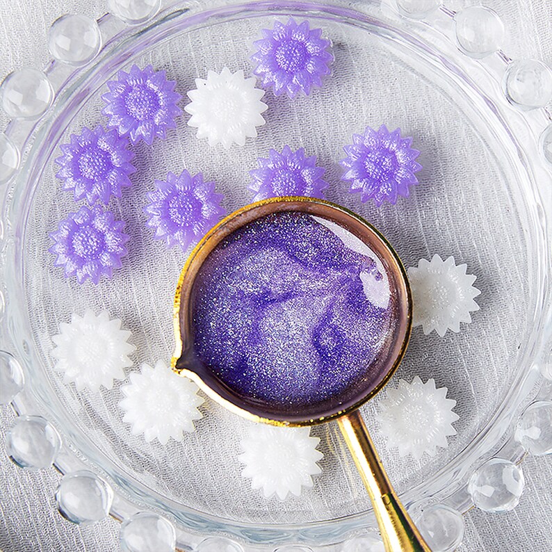 May include: A small glass dish with a gold rim contains a variety of purple and white flower-shaped resin embellishments. A small gold spoon with a purple glittery liquid sits in the center of the dish.