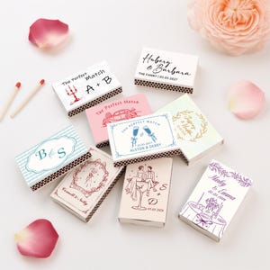 May include: A collection of rectangular matchboxes featuring various custom printed designs, including names, dates, and illustrations like champagne glasses, cars, and floral wreaths. The boxes have brown strike strips and are arranged with two loose matches.
