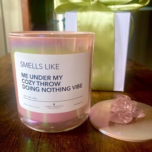 Op de afbeelding: Een roze iriserende glazen kaars met een wit label waarop staat "Smells Like Me Under My Cozy Throw Doing Nothing Vibe" en "Soy Luxe Wax". De kaars staat op een houten oppervlak met een roze deksel en een roze kristal op het deksel.