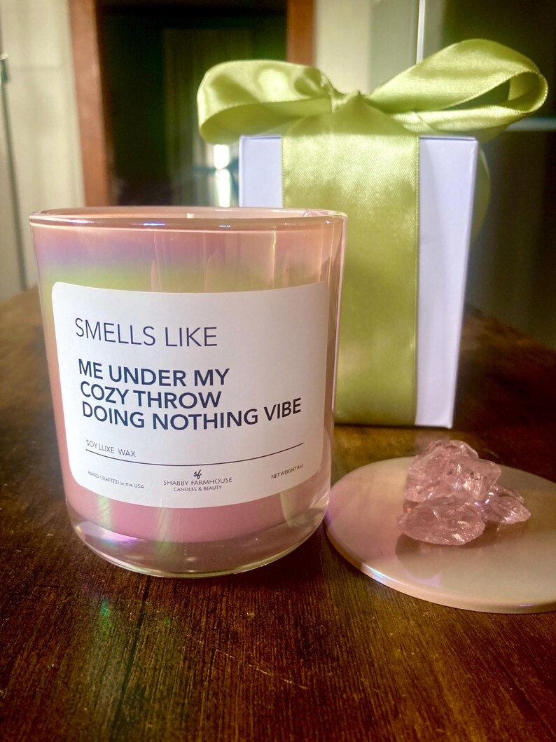 Op de afbeelding: Een roze iriserende glazen kaars met een wit label waarop staat "Smells Like Me Under My Cozy Throw Doing Nothing Vibe" en "Soy Luxe Wax". De kaars staat op een houten oppervlak met een roze deksel en een roze kristal op het deksel.