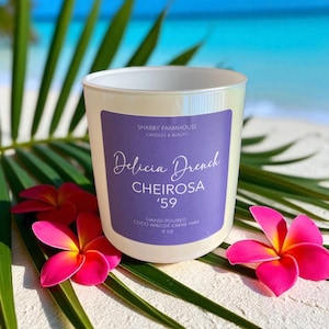 May include: A pearlescent candle with a purple label featuring the text "Delicia Drench Cheirosa '59". The candle is hand-poured with coco apricot creme wax and is 9 oz. Bright pink flowers and green leaves surround the candle.