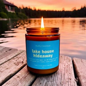 May include: A lit candle in a brown glass jar with a blue label that reads "lake house hideaway." The candle is on a wooden surface with a lake and sunset in the background. The candle is 8 oz.