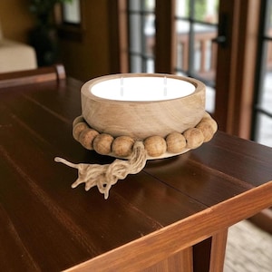 May include: A round, light-toned wooden bowl candle featuring three white wicks. The exterior of the bowl is encircled by a strand of natural wooden beads, with a decorative jute tassel hanging from the side of the bead arrangement.