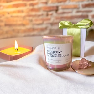 Op de afbeelding: Een roze iriserende glazen kaars met een wit label waarop staat "Smells Like Me Under My Cozy Throw Doing Nothing Vibe".