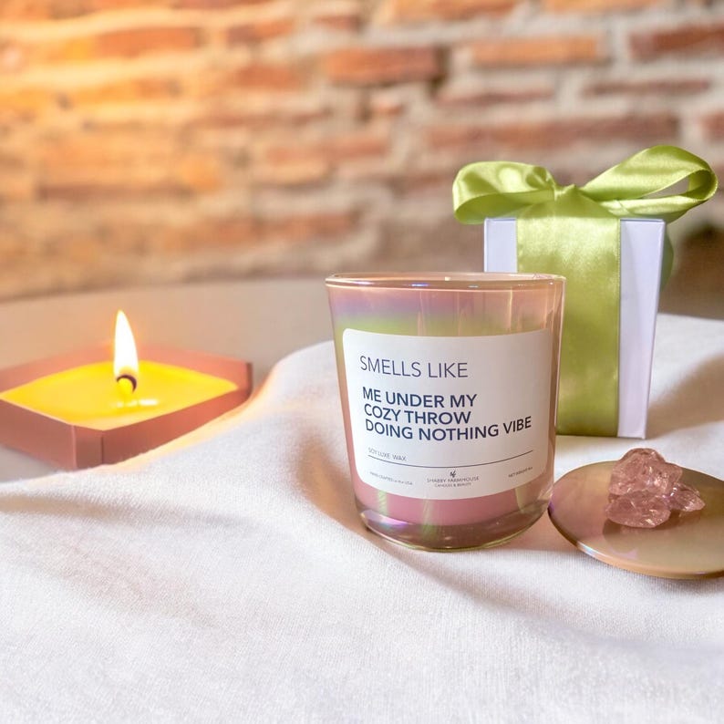 Op de afbeelding: Een roze iriserende glazen kaars met een wit label waarop staat "Smells Like Me Under My Cozy Throw Doing Nothing Vibe".