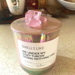 Op de afbeelding: Een roze iriserende glazen kaars pot met een wit label waarop staat "Smells Like Me Under My Cozy Throw Doing Nothing Vibe" Soy Luxe Wax. De kaars pot is bedekt met een roze kristal.