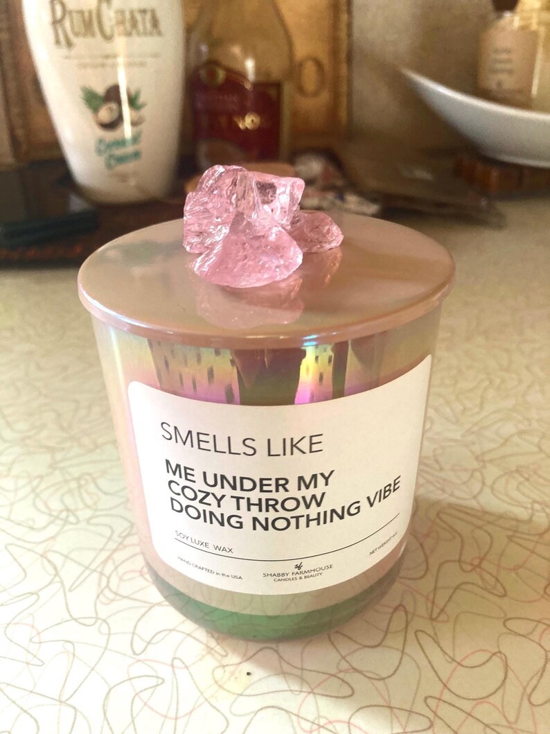 Op de afbeelding: Een roze iriserende glazen kaars pot met een wit label waarop staat "Smells Like Me Under My Cozy Throw Doing Nothing Vibe" Soy Luxe Wax. De kaars pot is bedekt met een roze kristal.