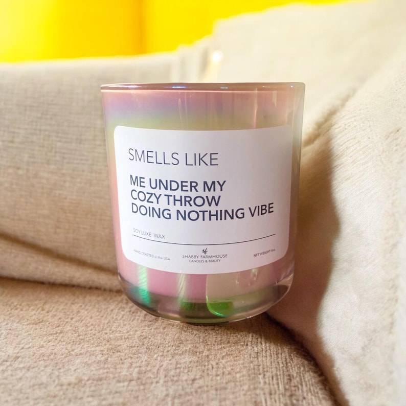 Op de afbeelding: Een roze iriserende glazen kaars met de tekst "SMELLS LIKE ME UNDER MY COZY THROW DOING NOTHING VIBE" en "SOY LIKE WAX" op het etiket. De kaars staat op een beige oppervlak.