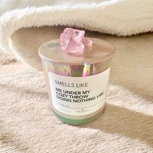 Op de afbeelding: Een roze iriserende kaars pot met een roze kristal erop. Het kaarsetiket luidt "Smells Like Me Under My Cozy Throw Doing Nothing Vibe" en "Soy Luxe Wax".