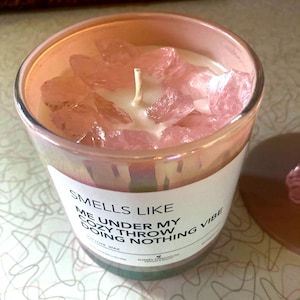 Op de afbeelding: Een roze kaars met een wit label waarop staat "Smells Like Me Under My Cozy Throw Doing Nothing Vibe" en is versierd met roze kristallen. De kaars is gemaakt van sojawas.