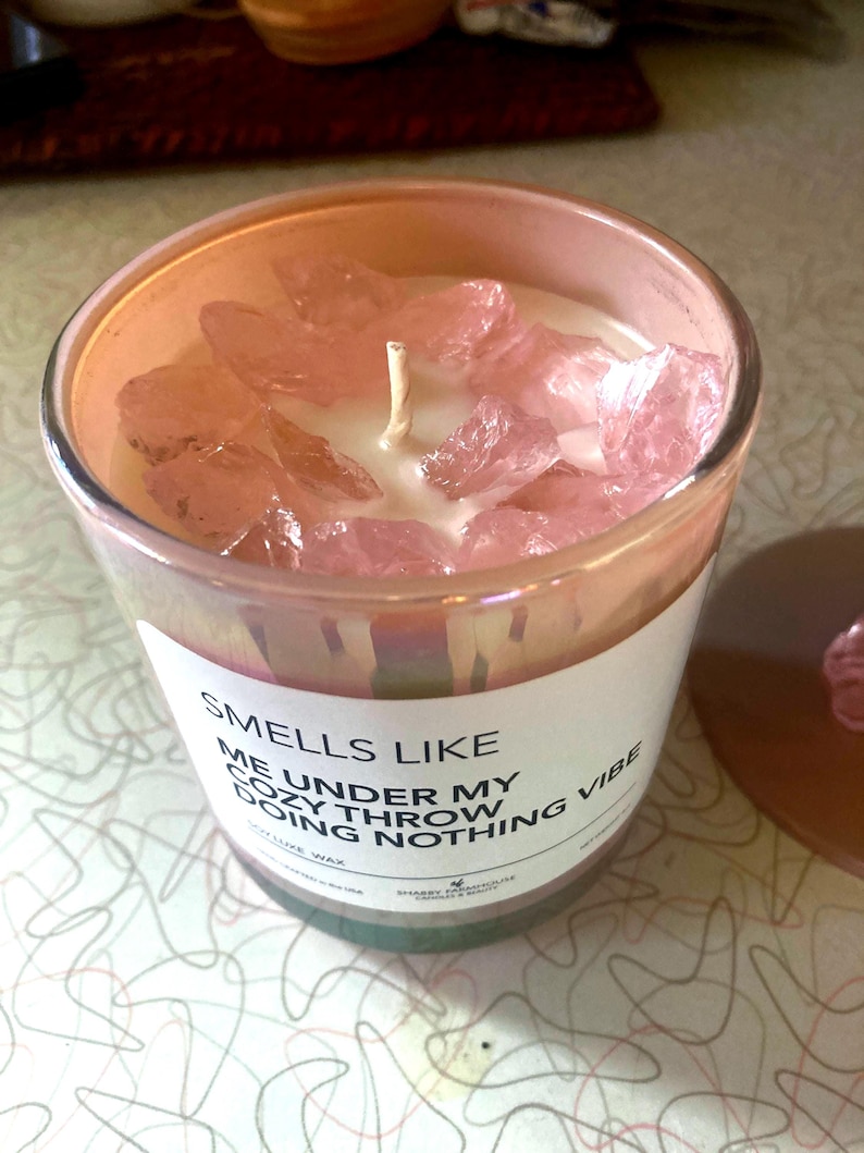 Op de afbeelding: Een roze kaars met een wit label waarop staat "Smells Like Me Under My Cozy Throw Doing Nothing Vibe" en is versierd met roze kristallen. De kaars is gemaakt van sojawas.