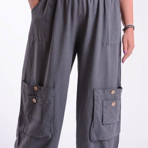 May include: Gray wide-leg pants with multiple pockets and wooden button details. The pants have an elastic waistband and a relaxed fit. The fabric appears to be a lightweight material, suitable for casual wear. The pants have two large pockets on the front.