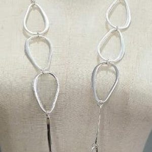 Puede incluir: Un collar plateado con un diseño abstracto. El collar presenta una cadena con eslabones ovalados y en forma de lágrima alargados. Los eslabones están conectados por barras rectangulares más pequeñas. El collar se exhibe en un maniquí.