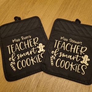 May include: Two black oven mitts with gold text that reads "Teacher of smart cookies" with a gingerbread man and candy cane graphic. The names "Miss Rivers" and "Miss Stewart" are printed on each mitt.