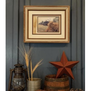 Puede incluir: Una impresión enmarcada de tractores y balas de heno vintage, con espigas de trigo. Una escena rústica con una estrella roja, una linterna y un pequeño barril de madera. La decoración está colocada contra una pared azul.
