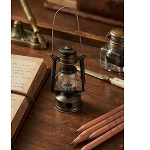 May include: A miniature metal lantern with a dark bronze finish, featuring a clear glass globe, a wire handle, and a circular base. The lantern is constructed with a central burner housing and a protective cage frame, standing upright as a decorative object.
