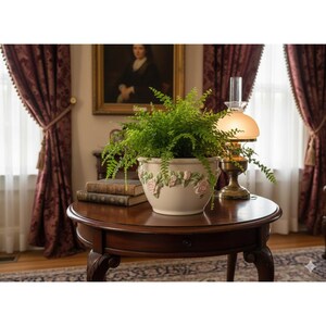 May include: A round, dark wood table with a fern in a cream-colored pot decorated with pink roses. A vintage lamp and stacked books sit nearby. The room has burgundy curtains and a painting on the wall.