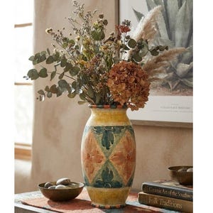 May include: A ceramic vase with a yellow base and a repeating diamond pattern in green and orange, filled with a bouquet of dried eucalyptus, pampas grass, and brown hydrangea blossoms. The vase sits beside a small bowl of smooth stones.