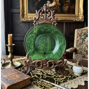 Puede incluir: Un plato de vidrio verde con un diseño floral en relieve se exhibe en un soporte de madera tallada. El plato es el punto focal, con una vela en un candelabro de latón y un libro antiguo. El diámetro del plato es de aproximadamente 25 cm.