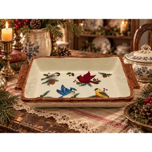 May include: A rectangular ceramic baking dish with a cream-colored interior featuring illustrations of a cardinal, a blue jay, a chickadee, and a goldfinch among pine branches and holly berries, framed by a textured brown faux-wood rim and handles.