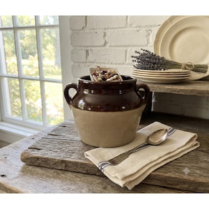 May include: A two-handled ceramic crock featuring a dark brown glazed upper section and a light tan glazed lower section. The crock is filled with a small square dish of mixed dried beans and sits on a wooden surface with a silver spoon and cloth.