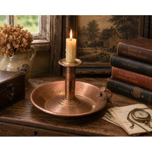 May include: A round copper candle holder with a central stem and a small side handle. A single cream-colored taper candle is inserted into the holder and is lit, producing a small flame. The copper surface shows a weathered, metallic patina.