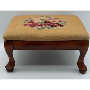 May include: A rectangular footstool with a tan needlepoint top featuring a central floral pattern in shades of pink, purple, and green. The base is constructed from polished dark brown wood and features four curved legs with rounded feet.