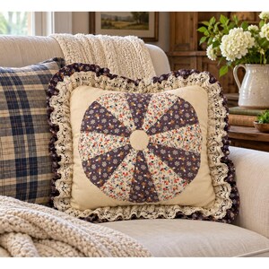 May include: A square throw pillow featuring a central circular patchwork design with alternating dark purple floral and cream floral fabric segments. The pillow is bordered by a dark purple ruffled edge trimmed with intricate cream lace.