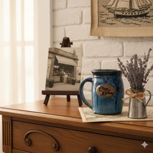 May include: A blue ceramic mug with a handle and the words "Origins of Brittany" on a wooden plaque. A small metal pitcher with lavender and a small framed picture are also on the wooden surface.