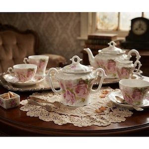 May include: A porcelain tea set featuring a white base with pink rose floral patterns. The set includes a teapot, a lidded sugar bowl with two handles, a creamer, three teacups with matching saucers, and a small square bowl with metal tongs.