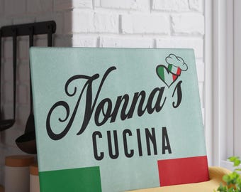 May include: A light blue cutting board with the text "Nonna's Cucina" in black cursive font. A red, white, and green heart with a chef's hat is above the text. The board has green and red stripes on the bottom.