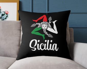 May include: Black cushion displaying the Trinacria symbol of Sicily, Italy, with red, green, and white details, and the word "Sicilia" in white script. The cushion is square and rests on a grey sofa.