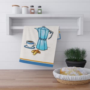 Op de afbeelding: Een blauwe en witte koffiepot met een kopje koffie en biscotti op een witte keukendoek met een gouden en blauwe rand.