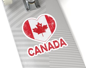 Op de afbeelding: Een rode sticker in de vorm van een hart met een verweerd Canadees vlagontwerp en het woord "CANADA" in witte letters. De sticker zit op een grijs-wit gestreept skateboard.