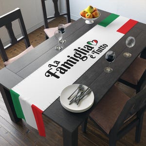 May include: White table runner with the text "La Famiglia e' Tutto" in black with a red heart. The runner has a green, white, and red Italian flag design on the edges.