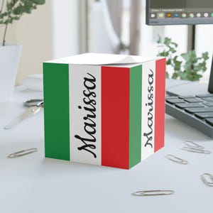 Puede incluir: Un bloc de notas cúbico con un diseño de rayas verdes, blancas y rojas. El nombre "Marissa" está escrito en cursiva en dos lados del cubo. El bloc de notas está sobre una superficie blanca con clips.