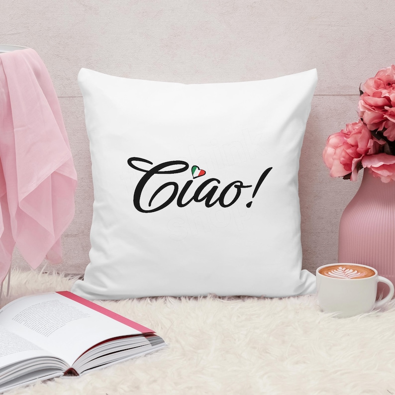 May include: White square pillow with the word "Ciao!" in black script, accented with a small Italian flag. The pillow is on a fluffy white surface, next to a pink vase with pink flowers and a cup of coffee.