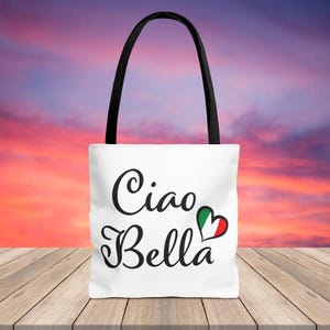 May include: White tote bag with black handles featuring the words "Ciao Bella" in elegant script. A heart-shaped graphic in the colors of the Italian flag is next to the words. The bag is on a wooden surface.