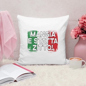 May include: White square pillow with the Italian flag colors and the phrase "MANGIA E STATTA ZITTO!" in black. The pillow is on a white fluffy surface, with a pink vase of flowers and a cup of coffee nearby.