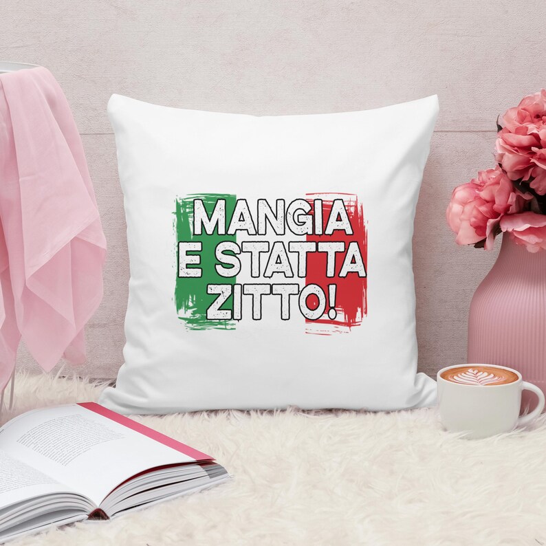 May include: White square pillow with the Italian flag colors and the phrase "MANGIA E STATTA ZITTO!" in black. The pillow is on a white fluffy surface, with a pink vase of flowers and a cup of coffee nearby.