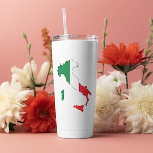 May include: White tumbler with a green, white, and red outline of Italy on it. The tumbler has a straw and is sitting on a pink surface with flowers.