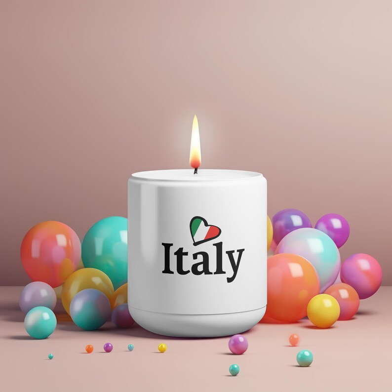 May include: A white candle with a burning flame, featuring the word "Italy" and a heart-shaped Italian flag design. The candle is surrounded by colorful spheres in various sizes and shades, including pink, orange, and blue.