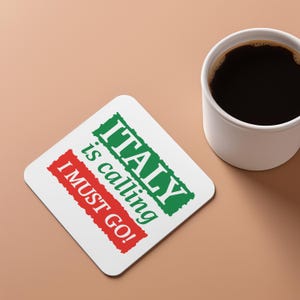以下が含まれることがあります： 「ITALY is calling I MUST GO!」の文字が緑と赤で書かれた白い正方形のコースター。濃いコーヒーが入った白いコーヒーマグが、コースターの隣のベージュ色の表面に置かれています。
