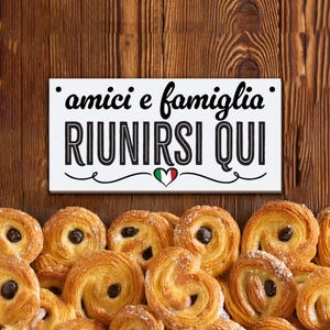以下が含まれることがあります： 中央に小さなイタリア国旗のハートマークが描かれた、「amici e famiglia riunirsi qui」という黒い文字が書かれた白い看板。看板は、たくさんのペストリーに囲まれている。
