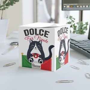 以下が含まれることがあります： コーヒーカップを持った白黒の猫のイラストが描かれた、白い立方体型のメモ帳。「DOLCE Far Niente」の文字が猫の上にあり、下に「Becca」という名前があります。イタリアの国旗が下にあります。