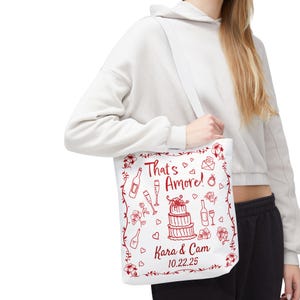 Puede incluir: Bolsa de lona blanca con un diseño rojo que presenta un pastel de bodas, botellas de vino y vasos. El texto dice "That's Amore! Kara & Cam 10.22.25".